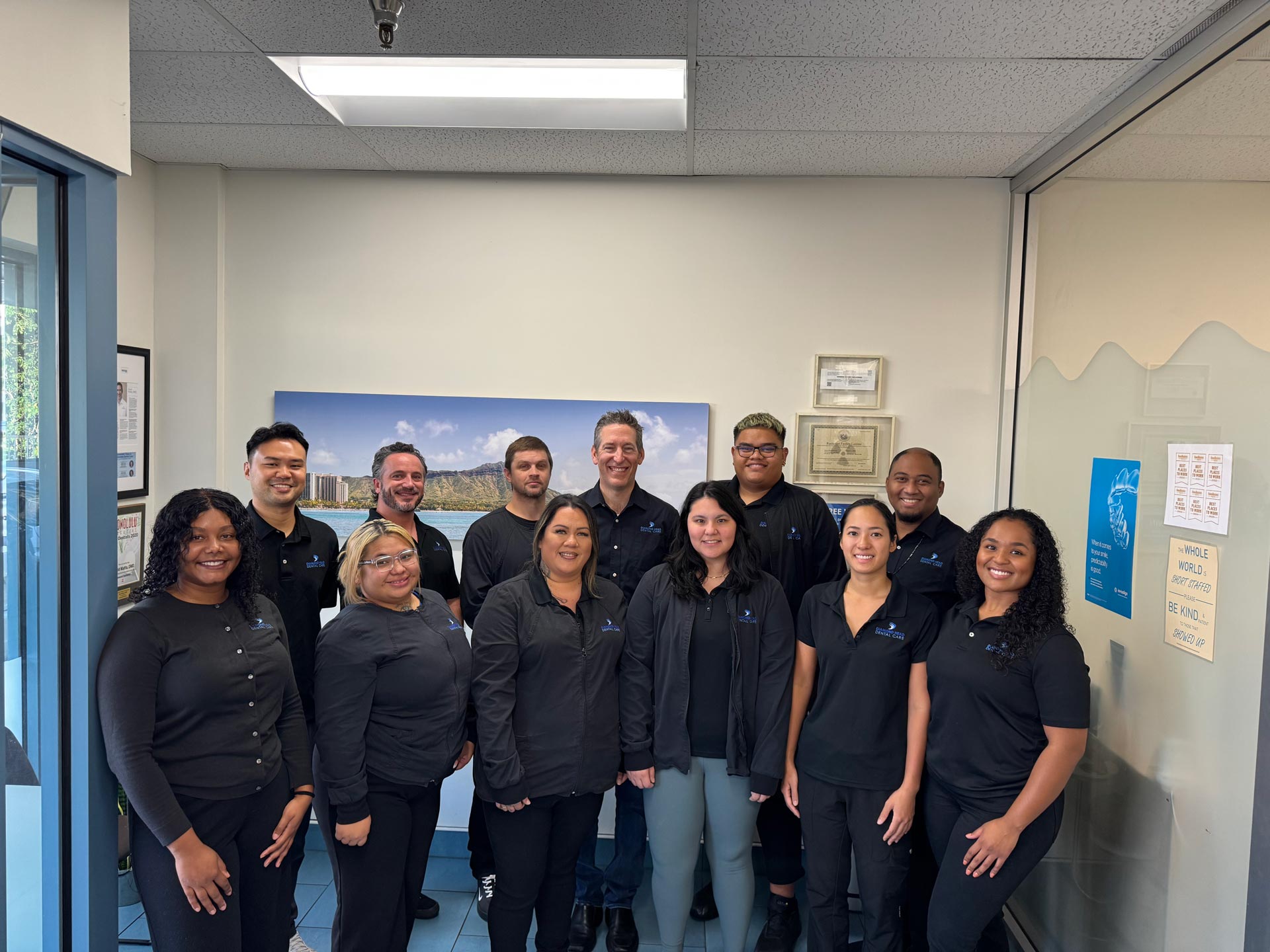 The image shows a group of people posing together in an office setting, with some individuals standing and others sitting behind them. They appear to be part of a professional team, possibly from a company, given the presence of a nameplate on the wall and a sign indicating a location or event.