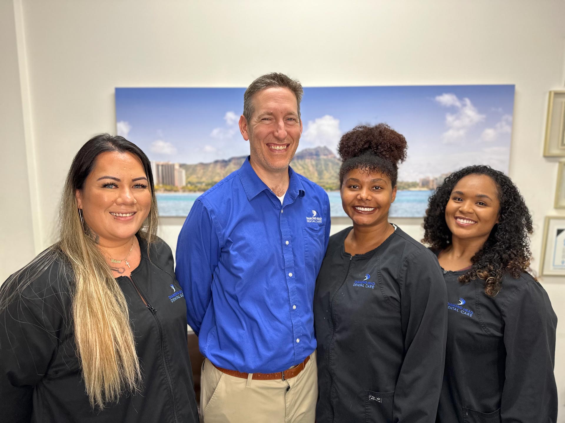 The image shows a group of people standing together, with a man on the left wearing a blue shirt and standing next to a large photographic print, and four women dressed in matching blue shirts with embroidered logos, suggesting they may be part of an organization or team.