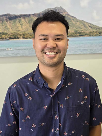 The image shows a person standing indoors with a smiling expression, wearing a patterned shirt, posing for a portrait-style photograph.
