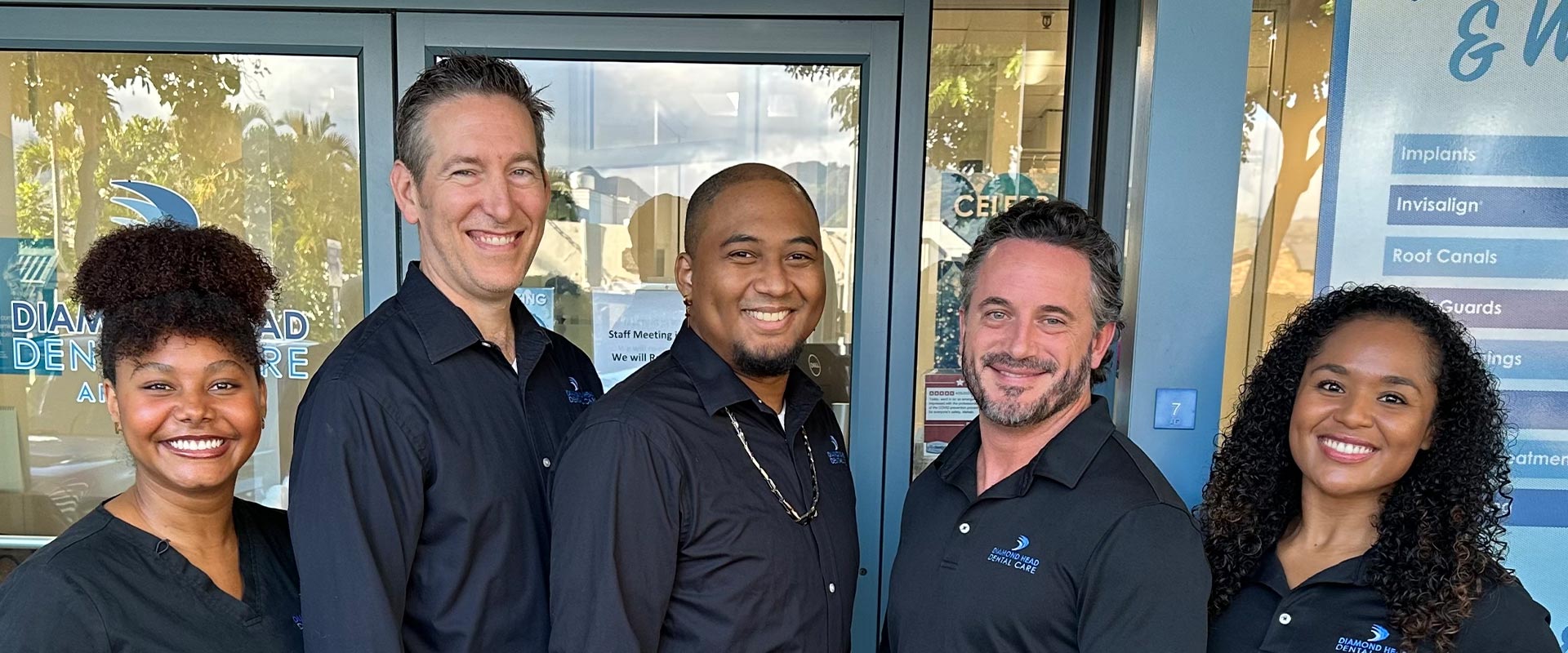A group of six individuals posing together, possibly for a promotional photo, with one person standing slightly behind the others, all smiling and appearing to be in front of an establishment with a sign that includes Sports and Cafe.