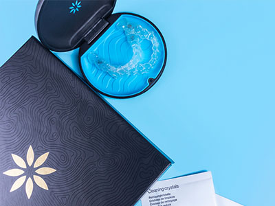 The image shows two open boxes with a blue interior, a small black object resembling a tea infuser, and a cup of water with a blue substance inside, all placed on a surface with a patterned background.