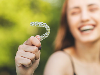 The image shows a person holding up a large toothbrush with a smiley face emoji on it against a blurred background.