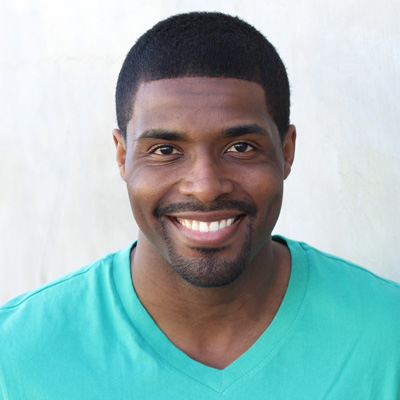 In the image, there are two older adults, a man and a woman, who appear to be enjoying each other s company. They are sitting close together, smiling, with the man on the left wearing a blue shirt and the woman on the right wearing a white top. The background suggests they might be in an outdoor setting, possibly near a body of water or a park, with greenery visible behind them.