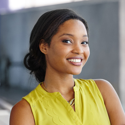 The image shows a woman smiling at the camera, wearing a yellow top and standing against a backdrop with a railing.