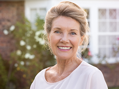 The image shows a woman with blonde hair smiling at the camera, wearing a white top, standing in front of a house with a brick wall and greenery.