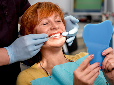 A woman sitting in a dental chair with a blue plastic mouthguard on her teeth, receiving dental treatment with a smiling expression, while holding a mirror up to her face.