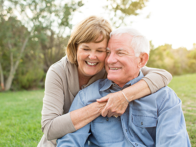 An elderly couple embracing each other outdoors during daylight, with both smiling and looking directly at the camera.