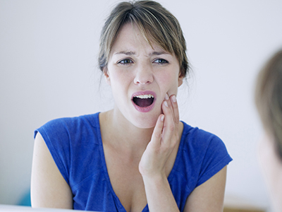 A woman with her mouth open, looking at her teeth, with a concerned expression, against a blurred background.