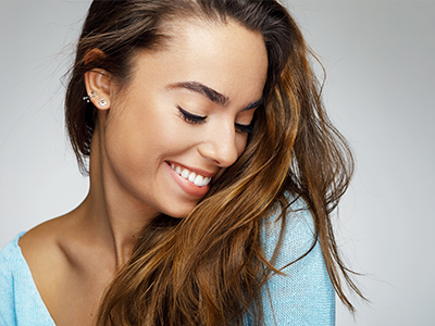 This image features a smiling woman with long hair, wearing a light blue top and earrings, looking over her shoulder while standing against a white background.