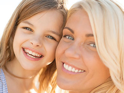 The image shows a woman with blonde hair smiling at the camera while holding a young girl who is also smiling. They appear to be outdoors, possibly on a beach, during daylight.