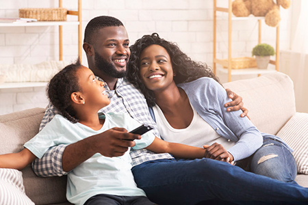 The image shows a family of four sitting on a couch, with a man holding a child and smiling at the camera, while another adult and a young child also look towards the camera. They appear to be enjoying a moment together in a cozy indoor setting.