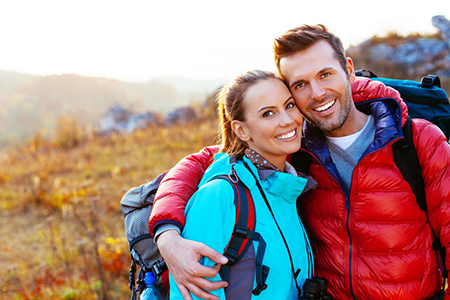 A man and woman posing together in a field, both wearing backpacks and smiling at the camera.