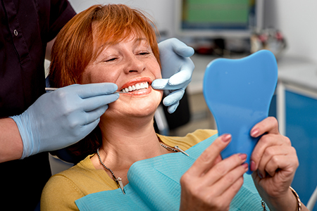 A woman with red hair wearing a blue surgical mask stands behind a dental hygienist who is holding up a tooth model for display, both are in a dental office setting.