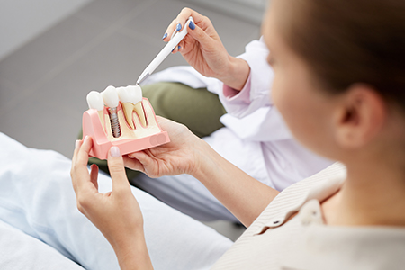 A dental professional holding a tooth model with a magnifying glass, demonstrating oral hygiene techniques to a patient lying on a hospital bed.