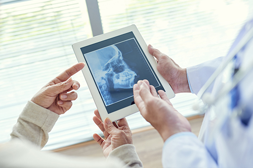 In the image, two individuals are engaged with a tablet displaying an X-ray, with one person holding the tablet and another showing the screen to them, both appear to be in a medical setting.