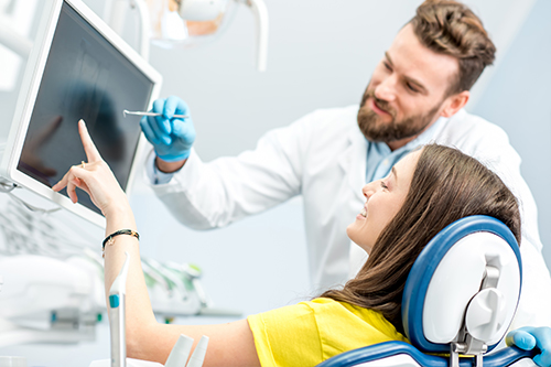 A man and woman in a medical setting, with the man standing behind a patient seated in a chair with an open mouth, examining teeth they are surrounded by dental equipment and technology.