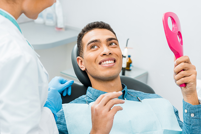 The image shows a person sitting in a dental chair with a smiling expression, holding a pink device in their hand, while receiving treatment from a dental professional who appears to be assisting them.
