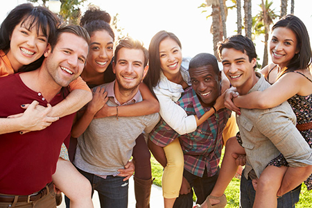 The image shows a group of young adults posing together outdoors, with some members hugging each other and smiling, under a bright sky during what appears to be sunset.